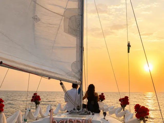 A couple seated at the bow under sail at sunset, with 'Happy Birthday' balloons and fresh roses